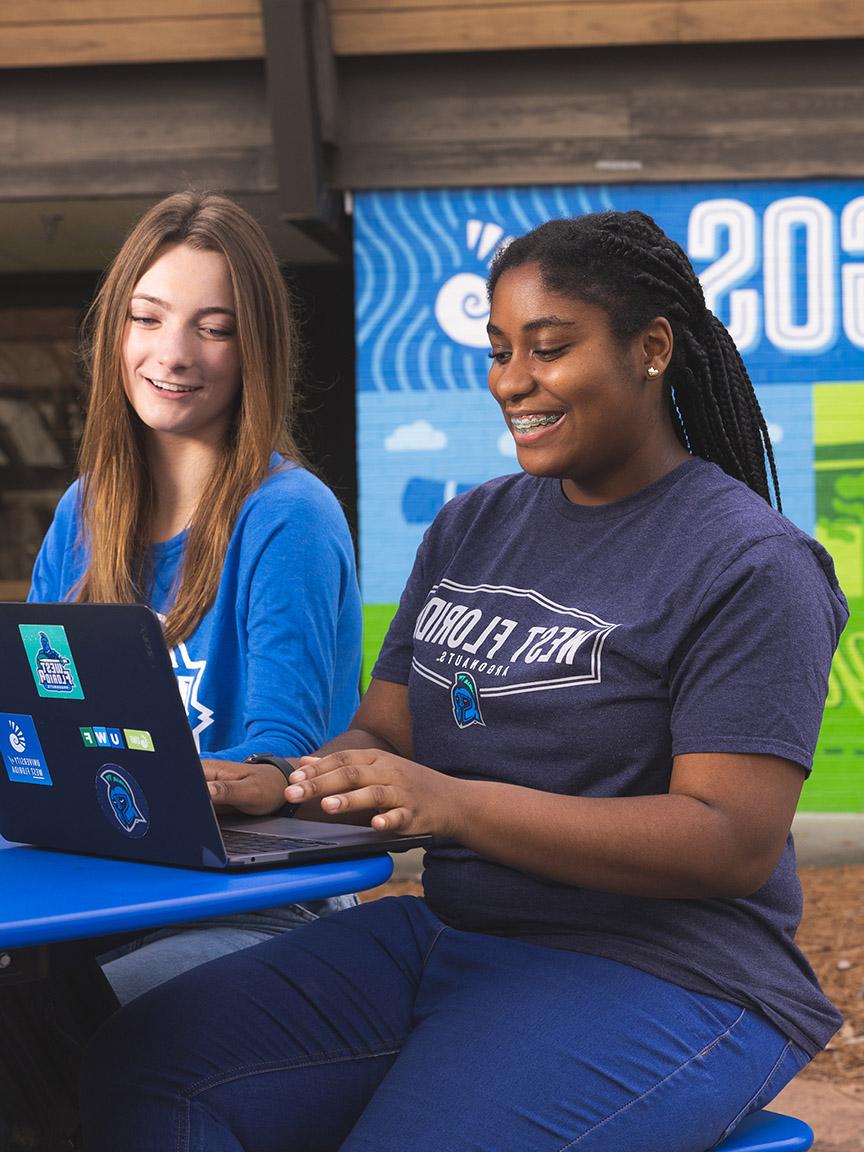 Two students smile while using laptops on the Commons patio near the UWF spirit mural.
